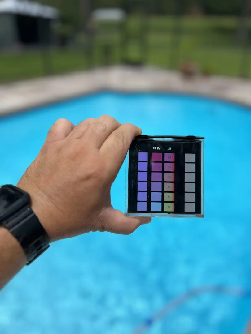 A hand holding a water test strip against its colour chart above a clear pool